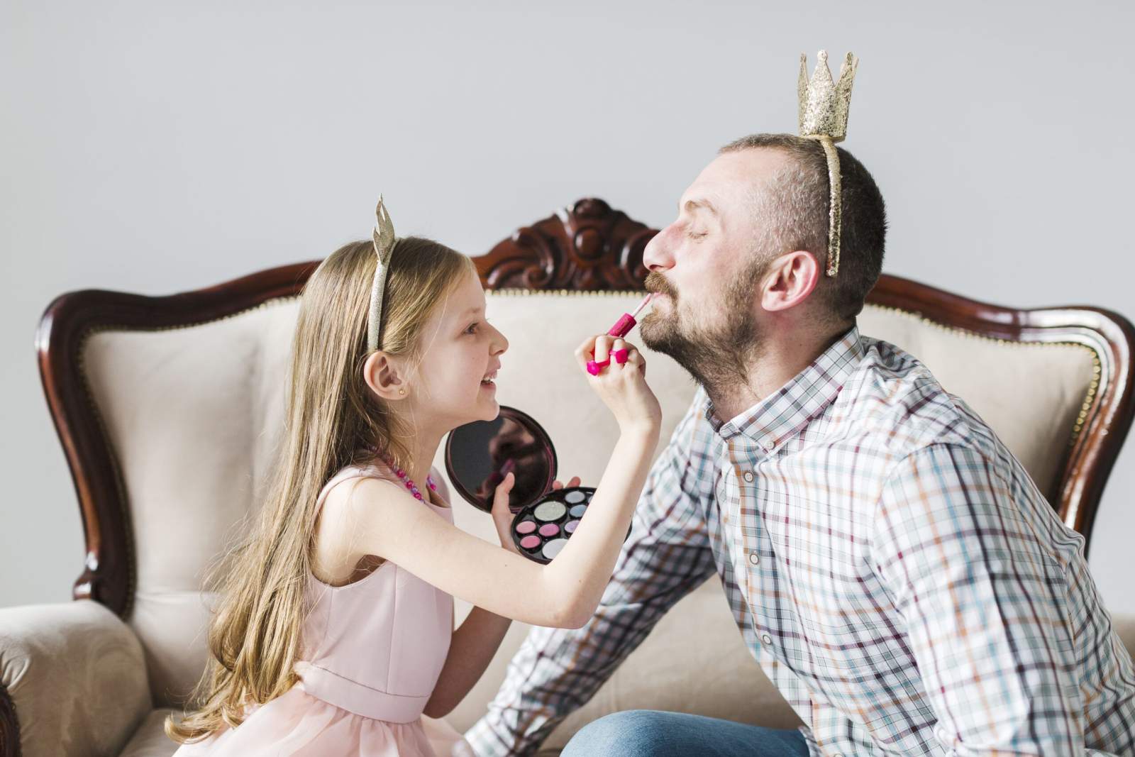 Vater und Tochter sitzen mit Krönchen auf einem Sofa. Der Vater wird dabei geschminkt.