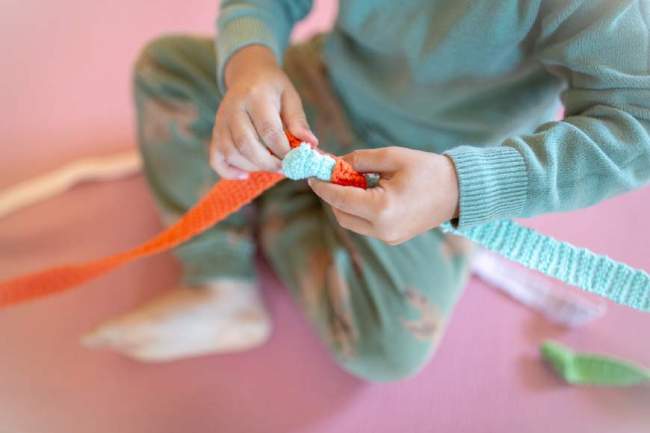 Kind sitzt auf dem Boden und verbindet bunte gehäkelte Streifen aus Wolle, Handarbeit und Kreativität beim Häkeln lernen.
