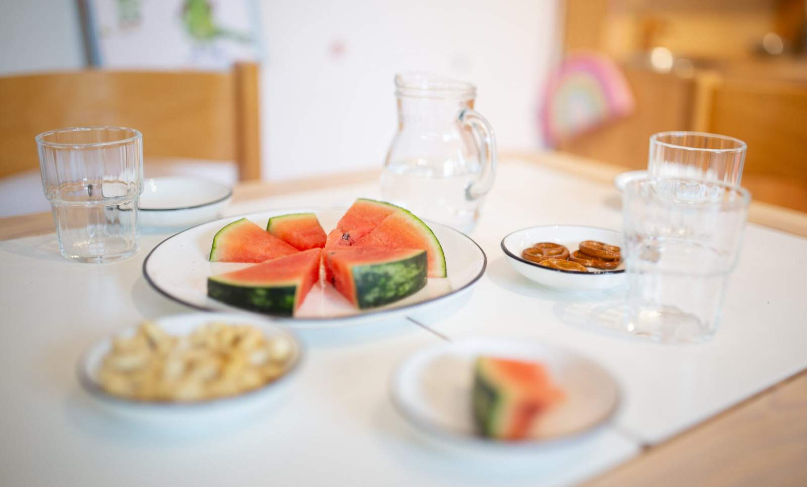 Gedeckter Tisch mit verschiedenen Snacks wie Wassermelone, Mini Brezeln und Nüssen, sowie eine Glaskanne Wasser und Gläser.